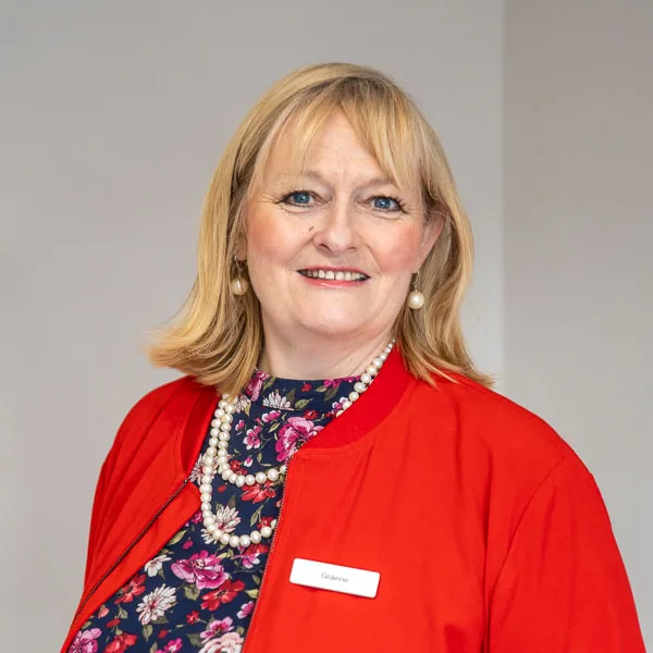 A smiling woman with blonde hair is wearing a red jacket over a floral top and pearl necklace. She is standing against a plain background.